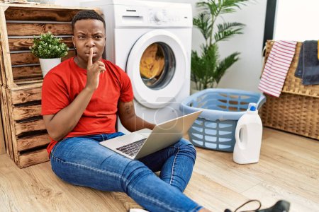 Téléchargez les photos : Jeune homme africain faisant la lessive et utilisant l'ordinateur demandant à être silencieux avec le doigt sur les lèvres. silence et concept secret. - en image libre de droit
