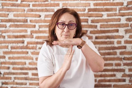 Photo for Senior woman with glasses standing over bricks wall doing time out gesture with hands, frustrated and serious face - Royalty Free Image