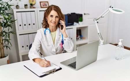 Foto de Mujer hispana de mediana edad vistiendo uniforme médico hablando en el teléfono inteligente en la clínica - Imagen libre de derechos
