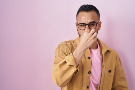 Foto de Young hispanic man standing over pink background smelling something stinky and disgusting, intolerable smell, holding breath with fingers on nose. bad smell - Imagen libre de derechos