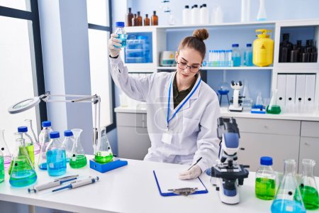 Photo for Young woman scientist writing on clipboard holding test tube at laboratory - Royalty Free Image