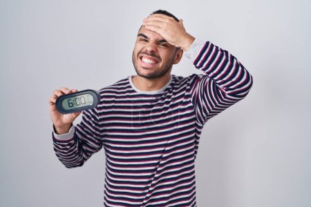 Photo for Young hispanic man wearing pyjama holding alarm clock stressed and frustrated with hand on head, surprised and angry face - Royalty Free Image