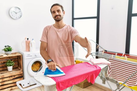 Foto de Joven hombre hispano planchando ropa en casa sonriendo amigable ofreciendo apretón de manos como saludo y bienvenida. negocios exitosos. - Imagen libre de derechos