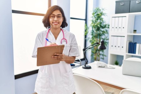 Photo for Young latin woman wearing doctor uniform writing on clipboard at clinic - Royalty Free Image