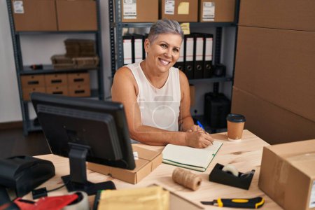 Téléchargez les photos : Femme d'âge moyen ecommerce travailleur d'affaires écrivant sur le livre au bureau - en image libre de droit