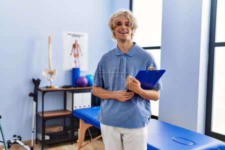 Téléchargez les photos : Jeune homme blond pysiothérapeute souriant confiant tenant le document à la clinique de réadaptation - en image libre de droit