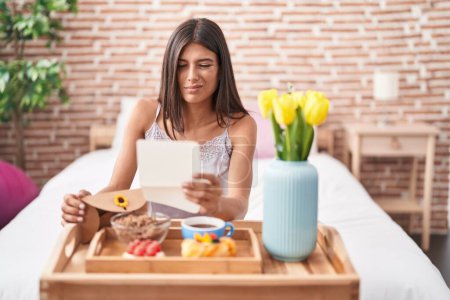 Foto de Morena mujer joven desayunando en la cama leyendo una carta despistada y confusa expresión. concepto de duda. - Imagen libre de derechos