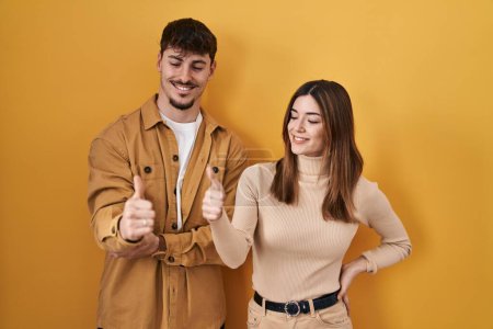 Téléchargez les photos : Young hispanic couple standing over yellow background looking proud, smiling doing thumbs up gesture to the side - en image libre de droit