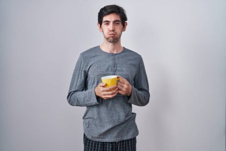 Téléchargez les photos : Young hispanic man wearing pajama drinking a cup of coffee puffing cheeks with funny face. mouth inflated with air, crazy expression. - en image libre de droit