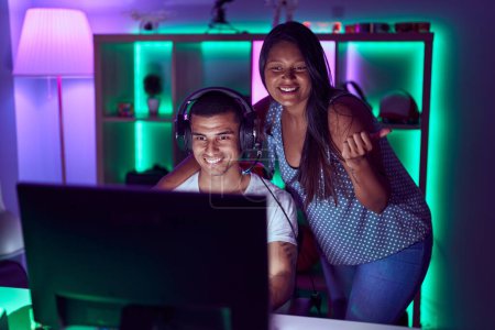 Foto de Joven pareja hispana jugando videojuegos apuntando con el pulgar hacia un lado sonriendo feliz con la boca abierta - Imagen libre de derechos