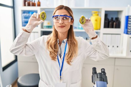 Téléchargez les photos : Jeune scientifique blonde femme travaillant avec de la nourriture au laboratoire souriant regardant vers le côté et regardant loin en pensant. - en image libre de droit