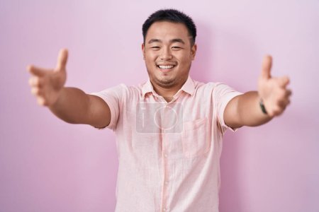 Téléchargez les photos : Jeune homme chinois debout sur fond rose regardant la caméra souriant à bras ouverts pour étreindre. expression joyeuse embrassant le bonheur. - en image libre de droit