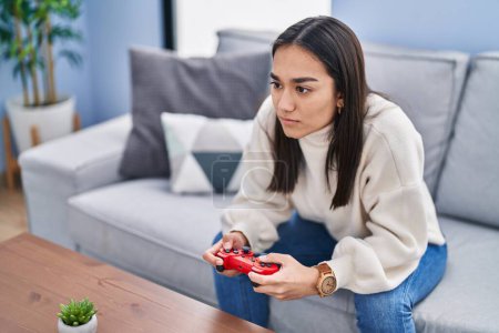 Photo for Young hispanic woman playing video game sitting on sofa at home - Royalty Free Image
