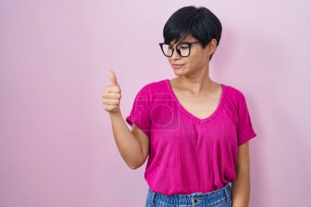 Photo for Young asian woman with short hair standing over pink background looking proud, smiling doing thumbs up gesture to the side - Royalty Free Image