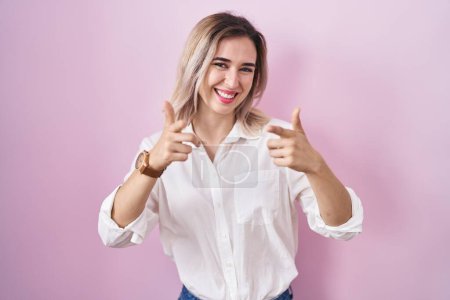 Foto de Young beautiful woman standing over pink background pointing fingers to camera with happy and funny face. good energy and vibes. - Imagen libre de derechos