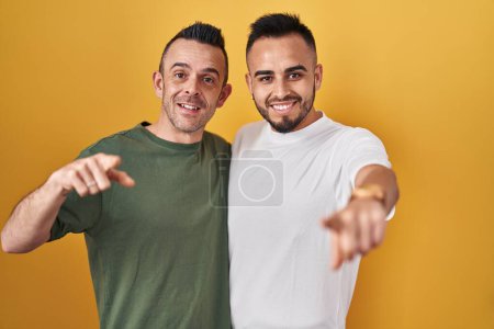 Photo for Homosexual couple standing over yellow background pointing to you and the camera with fingers, smiling positive and cheerful - Royalty Free Image