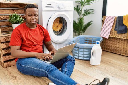 Foto de Young african man doing laundry and using computer depressed and worry for distress, crying angry and afraid. sad expression. - Imagen libre de derechos