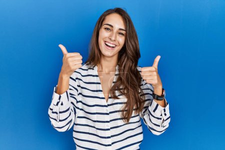 Foto de Young hispanic woman standing over blue isolated background success sign doing positive gesture with hand, thumbs up smiling and happy. cheerful expression and winner gesture. - Imagen libre de derechos