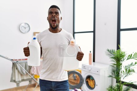 Photo for Young african man doing laundry holding detergent bottles angry and mad screaming frustrated and furious, shouting with anger looking up. - Royalty Free Image