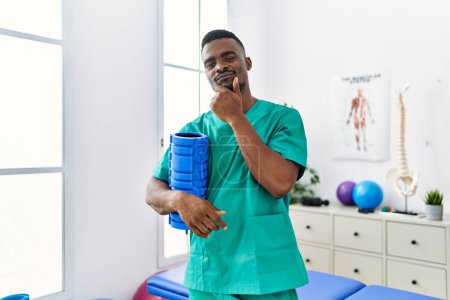 Foto de Young african physiotherapist man holding foam roller at the clinic serious face thinking about question with hand on chin, thoughtful about confusing idea - Imagen libre de derechos