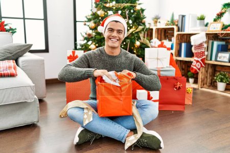 Téléchargez les photos : Jeune homme hispanique déballer cadeau assis par arbre de Noël à la maison - en image libre de droit