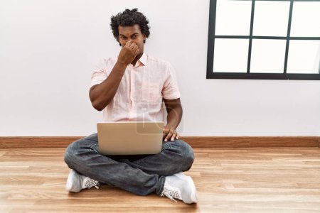 Téléchargez les photos : Homme africain avec les cheveux bouclés à l'aide d'un ordinateur portable assis sur le sol sentant quelque chose puant et dégoûtant, odeur intolérable, retenant souffle avec les doigts sur le nez. mauvaise odeur - en image libre de droit