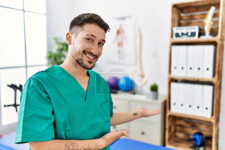 Foto de Joven fisioterapeuta hombre trabajando en la clínica de recuperación del dolor invitando a entrar sonriendo natural con la mano abierta - Imagen libre de derechos