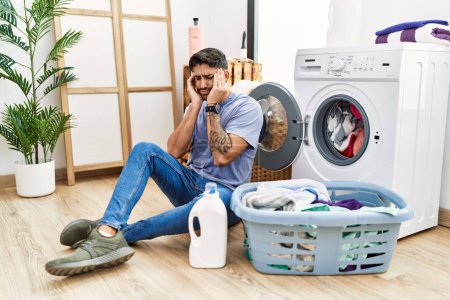 Téléchargez les photos : Young hispanic man putting dirty laundry into washing machine with hand on head, headache because stress. suffering migraine. - en image libre de droit