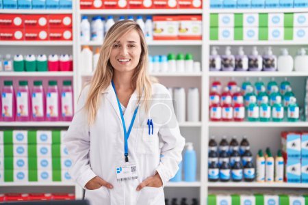 Foto de Joven mujer rubia farmacéutica sonriendo confiada de pie en la farmacia - Imagen libre de derechos