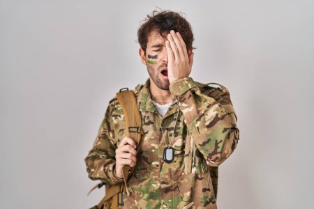 Foto de Joven hispano vistiendo uniforme camuflaje del ejército bostezando cansado cubriendo media cara, ojo y boca con la mano. cara duele en el dolor. - Imagen libre de derechos