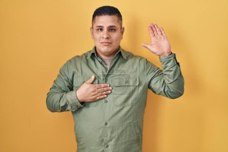 Téléchargez les photos : Jeune homme hispanique debout sur fond jaune jurer avec la main sur la poitrine et la paume ouverte, faire un serment de fidélité promesse - en image libre de droit