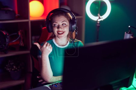Téléchargez les photos : Femme rousse jouant à des jeux vidéo souriant avec le visage heureux regardant et pointant vers le côté avec le pouce vers le haut. - en image libre de droit
