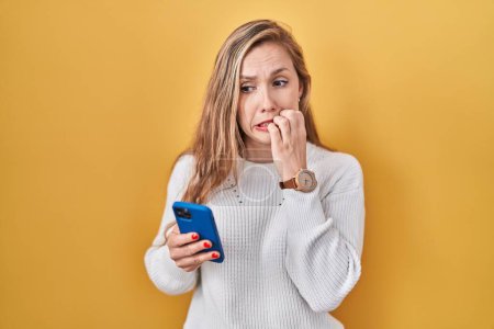 Téléchargez les photos : Jeune femme blonde utilisant le message de dactylographie smartphone regardant stressé et nerveux avec les mains sur les ongles mordants de la bouche. problème d'anxiété. - en image libre de droit