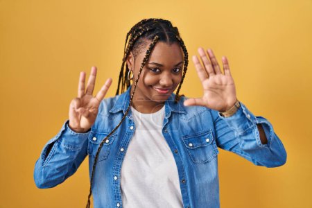Foto de Mujer afroamericana con trenzas de pie sobre fondo amarillo mostrando y apuntando hacia arriba con los dedos número ocho mientras sonríe confiado y feliz. - Imagen libre de derechos