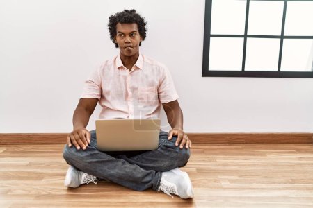 Téléchargez les photos : Homme africain avec les cheveux bouclés à l'aide d'un ordinateur portable assis sur le sol faisant visage de poisson avec des lèvres, geste fou et comique. expression drôle. - en image libre de droit