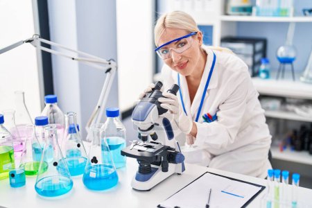 Téléchargez les photos : Jeune femme blonde portant un uniforme de scientifique au microscope au laboratoire - en image libre de droit