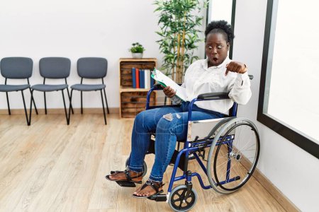 Téléchargez les photos : Jeune femme noire assise en fauteuil roulant dans la salle d'attente pointant vers le bas avec les doigts montrant la publicité, visage surpris et bouche ouverte - en image libre de droit
