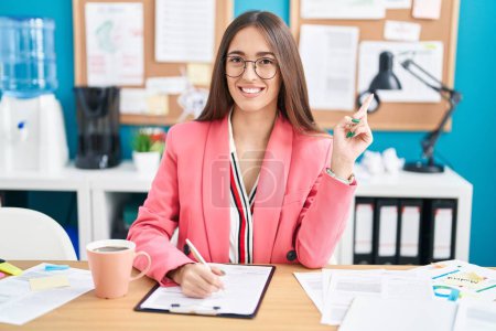 Téléchargez les photos : Jeune femme hispanique travaillant au bureau portant des lunettes gaies avec un sourire sur le visage pointant avec la main et le doigt sur le côté avec une expression heureuse et naturelle - en image libre de droit