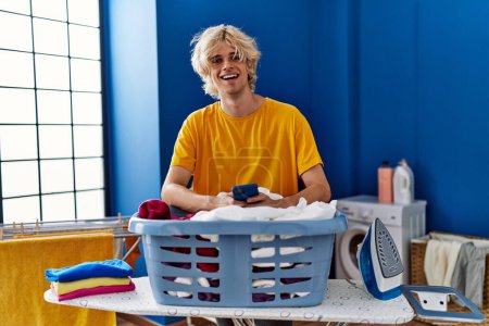 Photo for Young blond man using smartphone leaning on basket with clothes at laundry room - Royalty Free Image