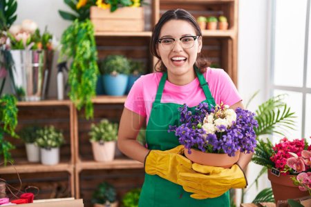 Photo for Hispanic young woman working at florist shop holding plant smiling and laughing hard out loud because funny crazy joke. - Royalty Free Image