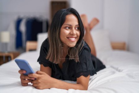 Photo for Young hispanic woman using smartphone lying on bed at bedroom - Royalty Free Image