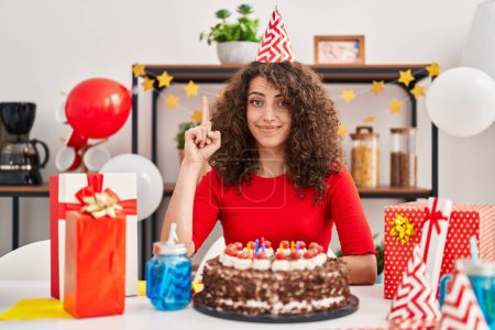 Foto de Hispanic woman with curly hair celebrating birthday holding big chocolate cake surprised with an idea or question pointing finger with happy face, number one - Imagen libre de derechos