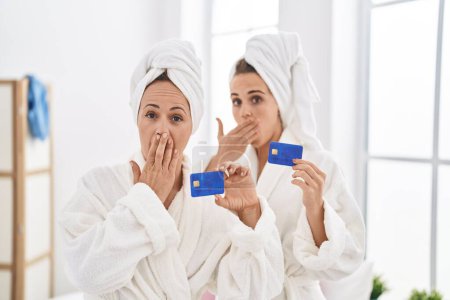Téléchargez les photos : Middle age woman and daughter wearing bathrobe holding credit card covering mouth with hand, shocked and afraid for mistake. surprised expression - en image libre de droit