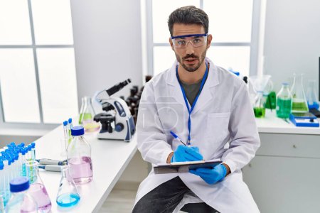Photo for Young hispanic man wearing scientist uniform writing on clipboard working at laboratory - Royalty Free Image