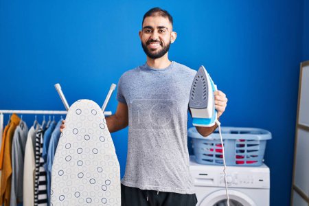 Téléchargez les photos : Middle east man with beard holding electric iron smiling with a happy and cool smile on face. showing teeth. - en image libre de droit