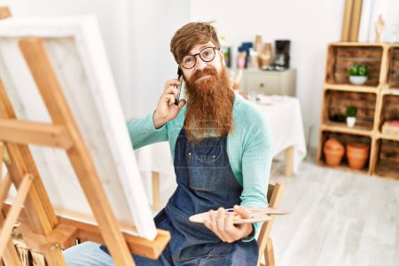Téléchargez les photos : Jeune rousse homme souriant dessin confiant et parler sur le smartphone au studio d'art - en image libre de droit