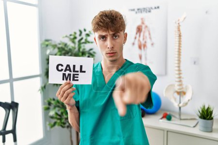 Téléchargez les photos : Young caucasian physio man holding call me banner at the clinic pointing with finger to the camera and to you, confident gesture looking serious - en image libre de droit