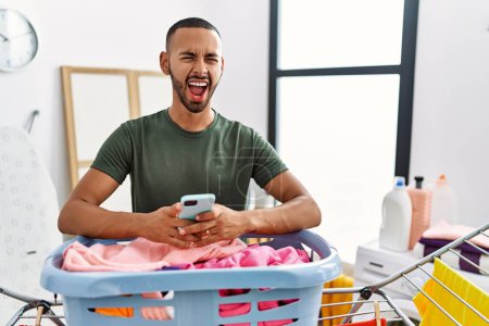 Foto de African american man doing laundry using smartphone angry and mad screaming frustrated and furious, shouting with anger. rage and aggressive concept. - Imagen libre de derechos