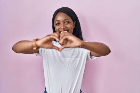 Photo for African young woman wearing casual white t shirt smiling in love doing heart symbol shape with hands. romantic concept. - Royalty Free Image