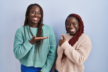 Photo for Two african woman standing over blue background showing palm hand and doing ok gesture with thumbs up, smiling happy and cheerful - Royalty Free Image